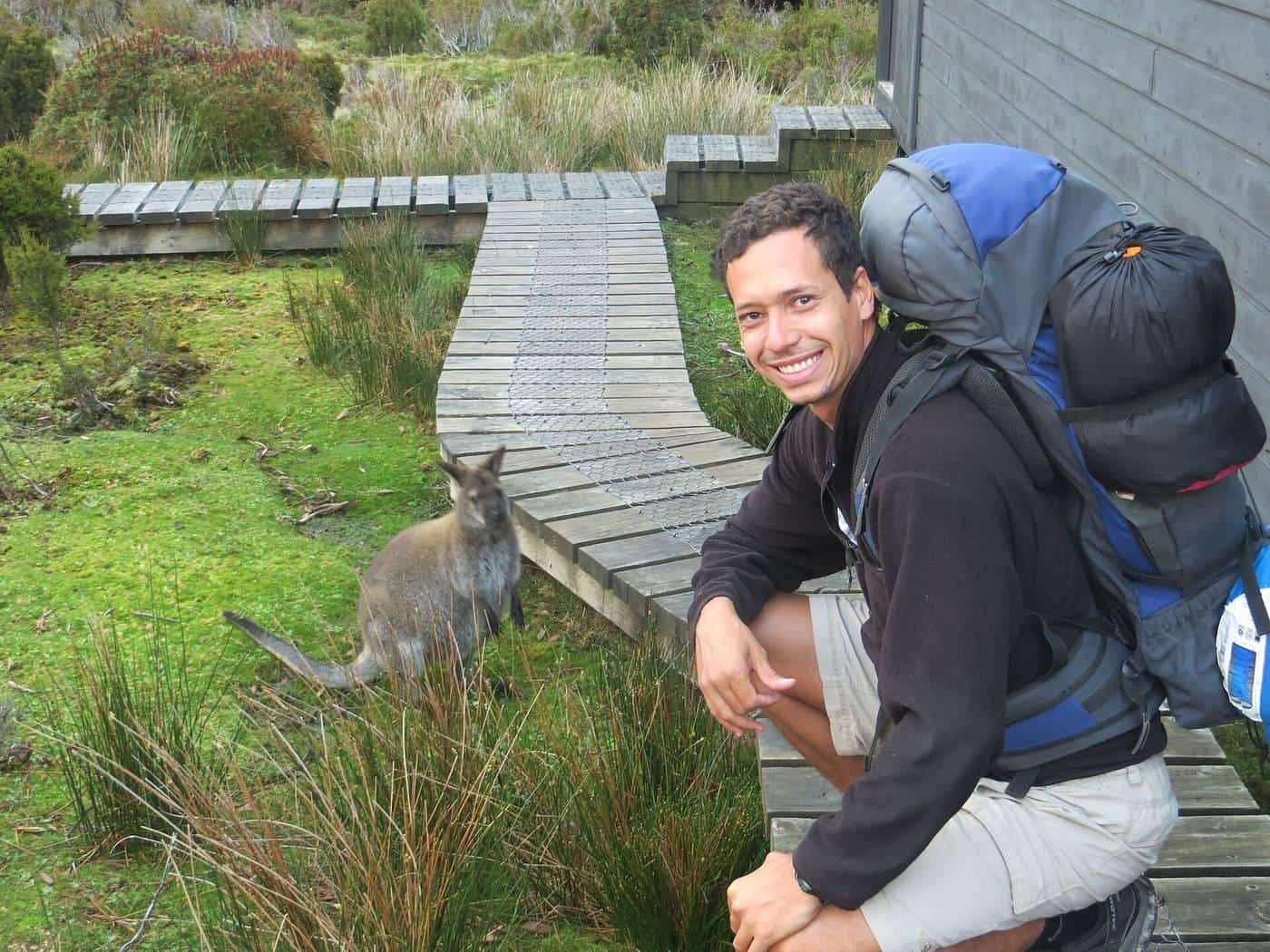 A smiling hiker with a large pack beside a boardwalk, with a wallaby nearby